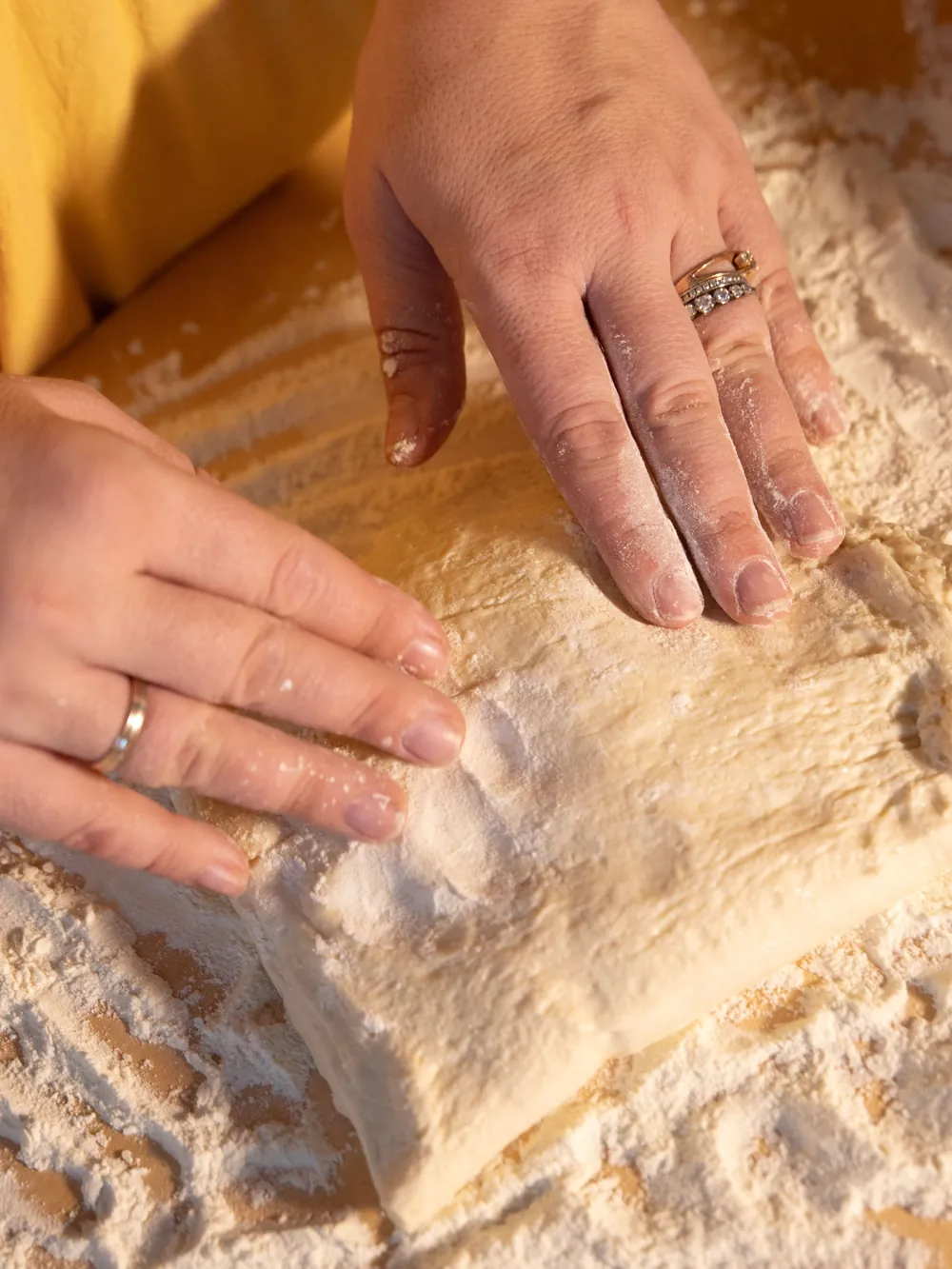 Dvě ruce poprášené moukou vtlačují kulatý plát pizza těsta na dřevěné desce