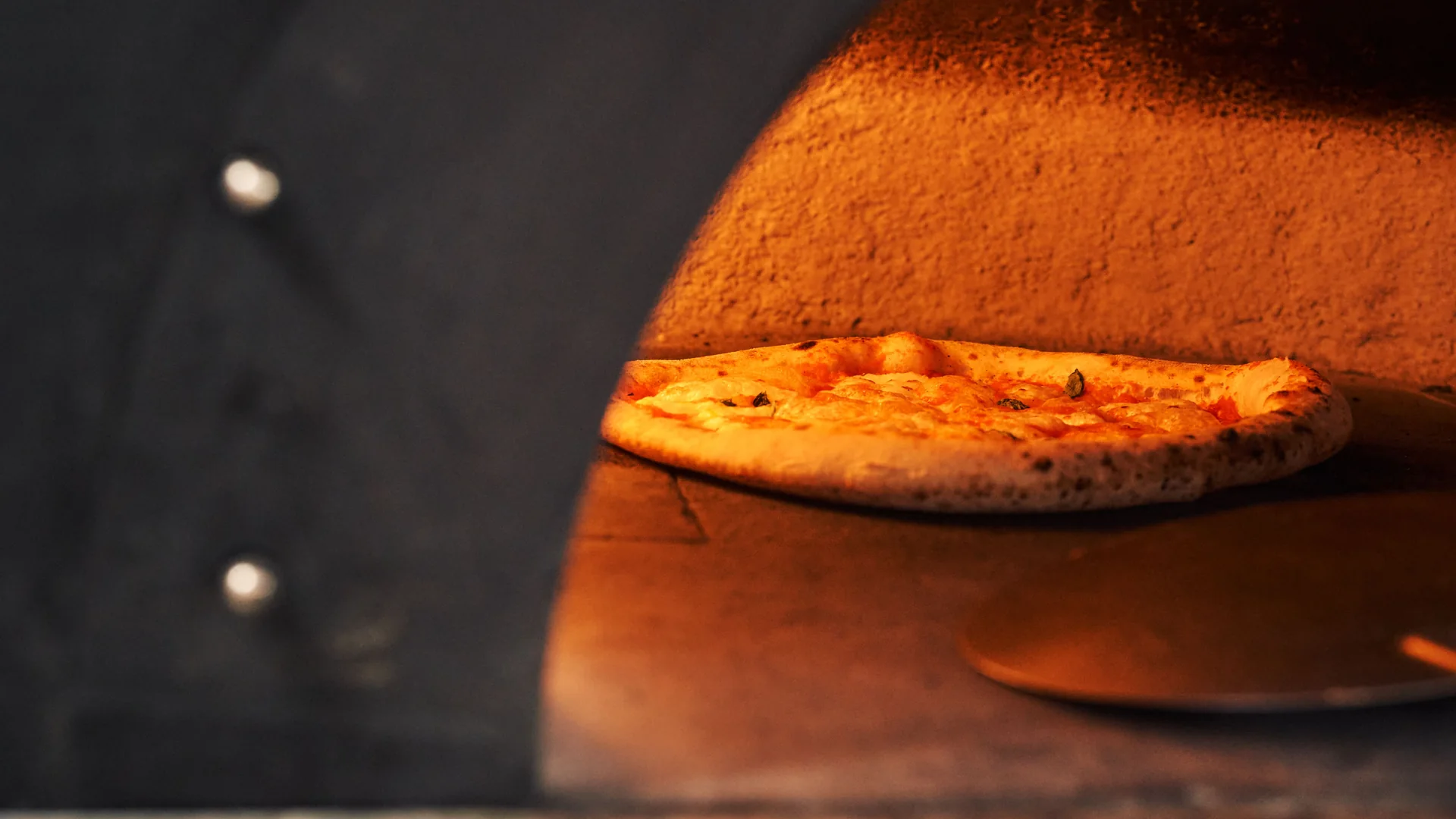 Pizza s puchýřnatou, očazenou kůrkou odpočívající na dně rozžhavené pece na dřevo, vedle ní leží kulatá kovová lopata