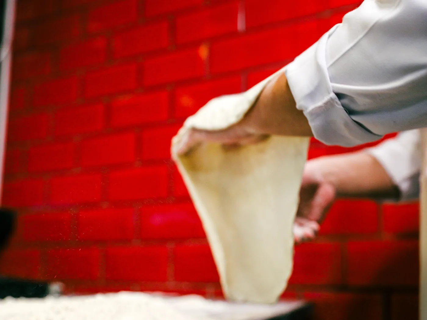 Kuchař v bílé bundě zvedá roztažený kulatý plát pizza těsta před cihlovou zdí